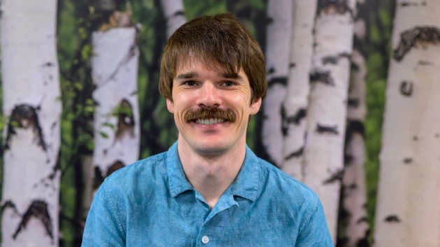 En man med mustasch och blå skjorta poserar framför en bakgrund med björkar