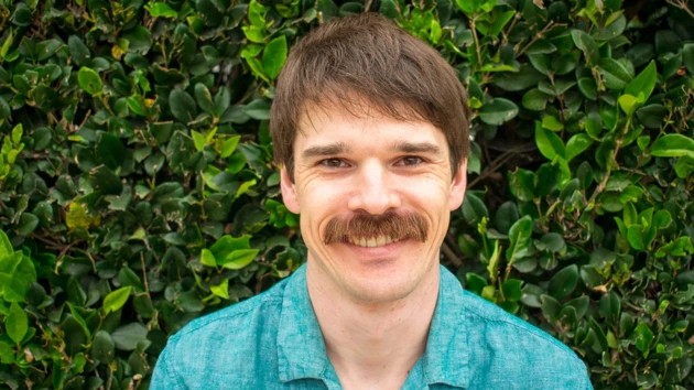 A man, wearing a blue shirt, with a rich moustache and smile on his
face is standing in front of a thick, bright green hedge.