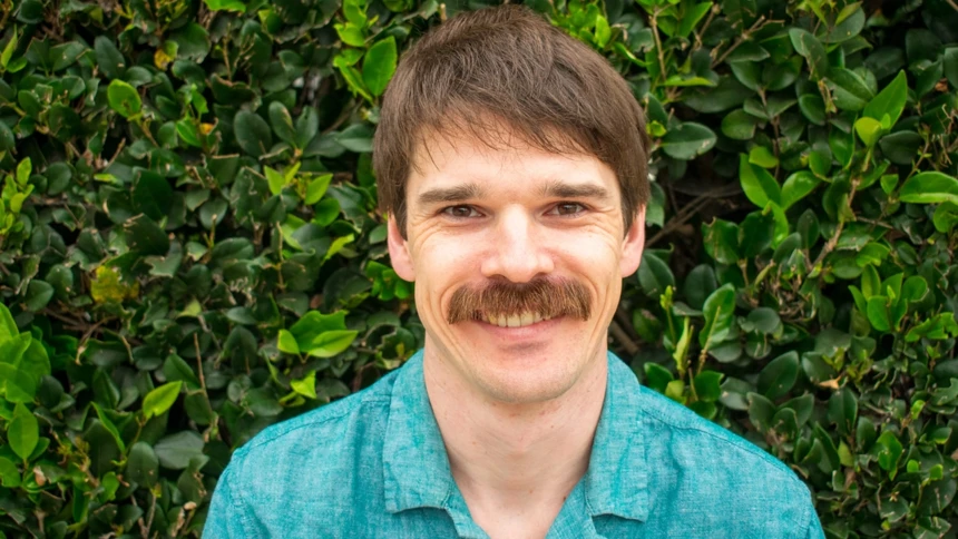 A man, wearing a blue shirt, with a rich moustache and smile on his
face is standing in front of a thick, bright green hedge.