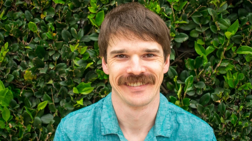 A man, wearing a blue shirt, with a rich moustache and smile on his
face is standing in front of a thick, bright green hedge.