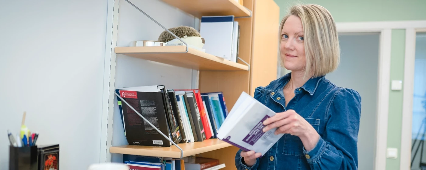 A woman is stood by a bookshelf browsing a book.