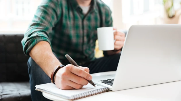 Person sitting on a sofa working. Laptop, coffee and notepad in front of him.