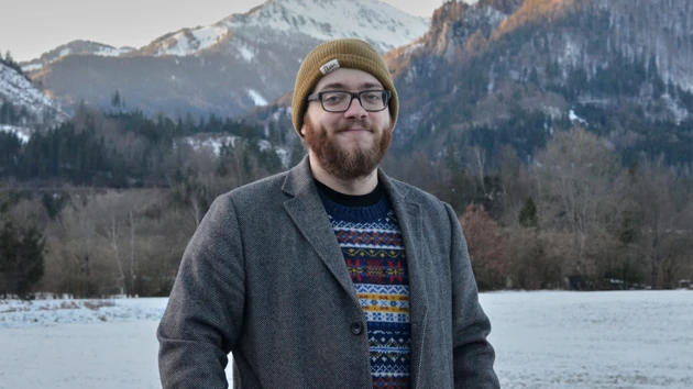 Bild på Bastian Schiffthaler stående utomhus, i bakgrunden sjö, skog och snöklädda berg