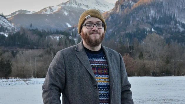 Photo of Bastian Schiffthaler standing outdoors, in the background lake, forest and snowcovered mountain tops