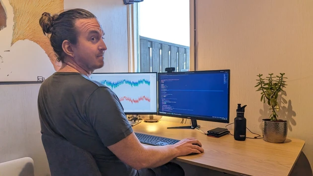 Bild på Daniel Dahlstrand sedd snett bakifrån, stående vid skrivbord med två datorskärmar