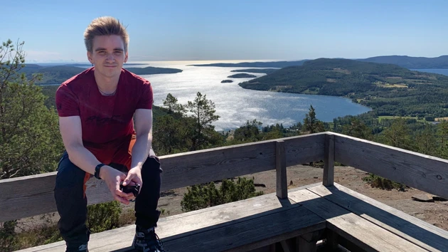 Bild på Fredrik Bredberg, sittande på ryggstödet till en bänk, i bakgrunden havsvik och berg