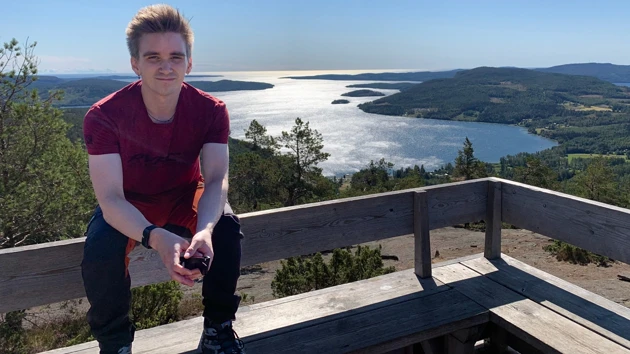 Bild på Fredrik Bredberg, sittande på ryggstödet till en bänk, i bakgrunden havsvik och berg