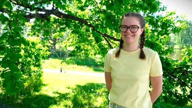 Hedvig Lind står under ett träd, i gul tröja och glasögon.