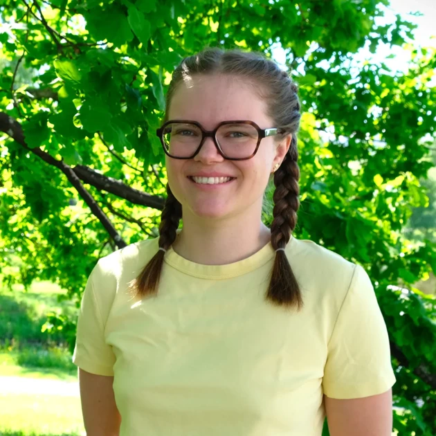 Hedvig Lind står under ett träd, i gul tröja och glasögon.