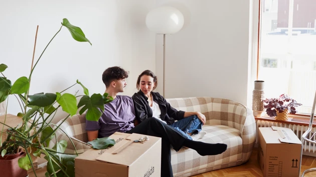 En man och en kvinna sitter i en soffa i ett vardagsrum, omgivna av flyttkartonger. 