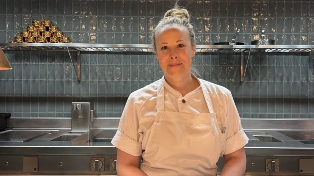 Emma Wixner, står i ett restaurangkök med kökskläder i vitt och grönt kakel i bakgrunden. Samt spis och hyllor i aluminium syns i bakgrunden.