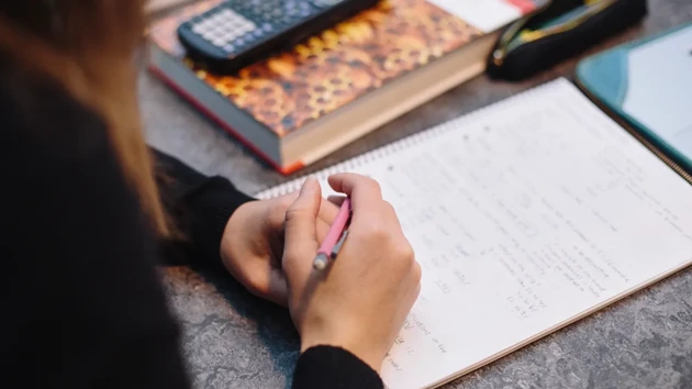 Hand writing on a notepad with a book and a calculator showing.
