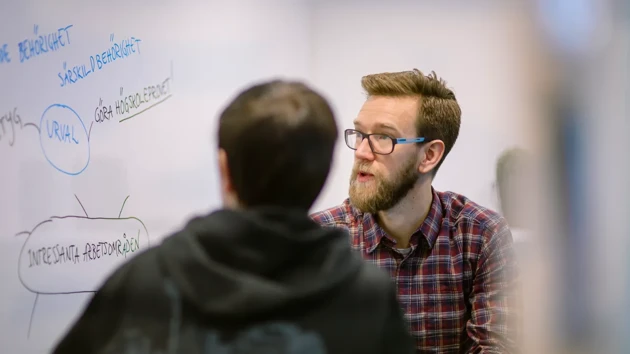 En man tittar på en whiteboard samtidigt som han samtalar med en annan person.