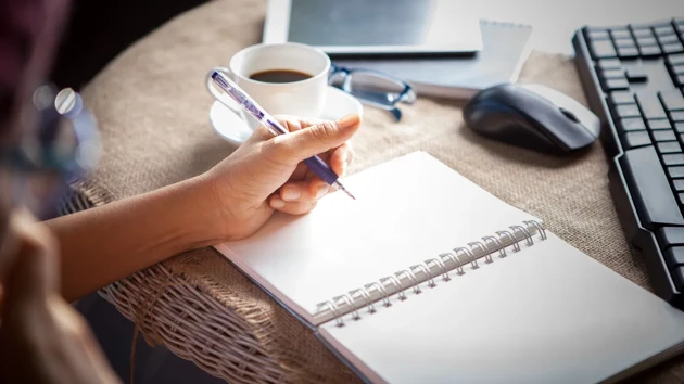 A person sitting at a desk prepared to take notes, using pen and paper. A computer is noticeable in the background.