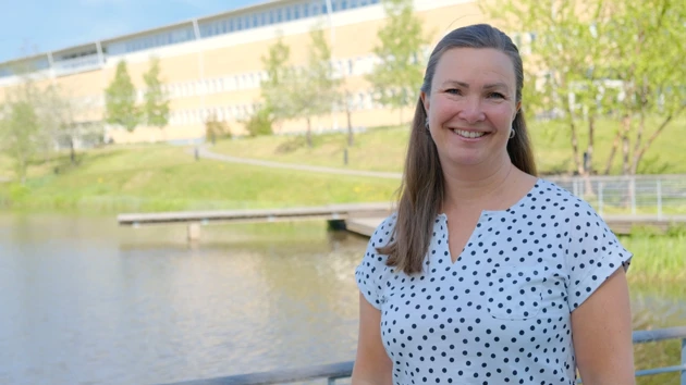 Karin Åström is standing in front of the Social science building by the pond. 