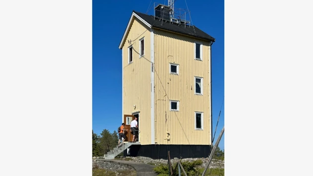 A wooden building in 3 stories, people going into the entrance door on a staircase. 