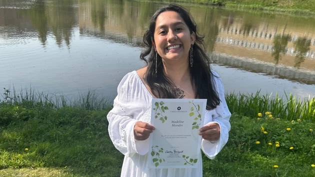 Foto av Madeline Morales hållande sitt masterdiplom, i bakgrunden campusdammen
