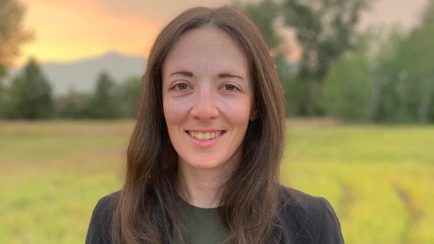 A woman with long dark hair, wearing dark casually elegant clothes, is
standing in an open field during a colourful sunset.