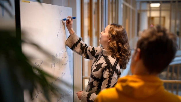 Kvinna skriver formler på whiteboard medan en man tittar på.