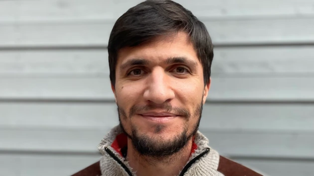 Porträtt på Najmaalddin Saifaddin Dawoud stående mot en väggpanel i grått trä.