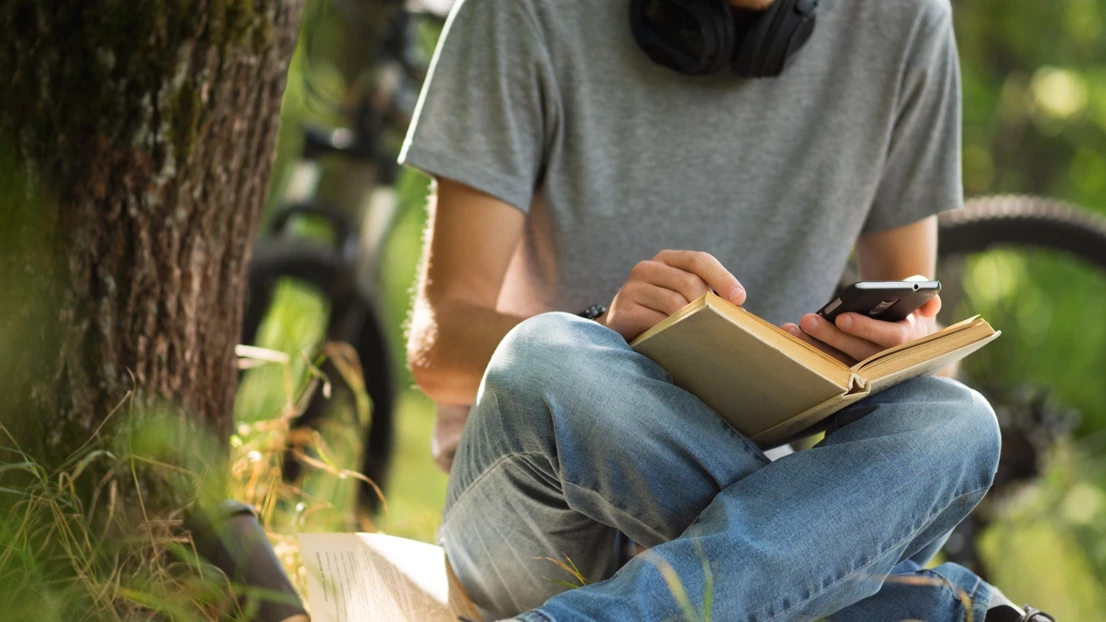 Kille som sitter utomhus i gräset vid ett träd med en mobil och en bok.