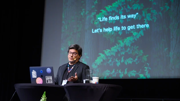 A man with dark hair and glasses standing at a table with a laptop in front of him and a screen behind. 
