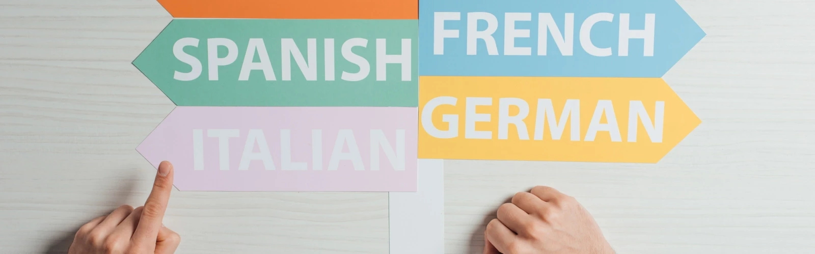 Skyltar med olika språk på som pekar åt olika håll som en hand pekar på. För att illustrera språkinlärning eller livslångt lärande.