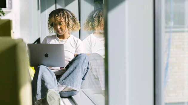 En student sitter i en solig fönstersmyg och läser på en laptop. 