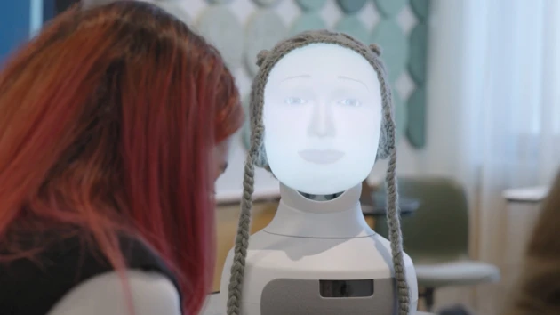 Foto av ett robotansikte med två människor som skymtar i förgrunden