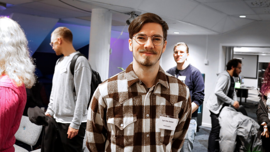 Bilden visar en manlig student, med mörkt hår och glasögon som tittar in i kameran. I bakgrunden syns andra studenter som deltog på Hackatonet.