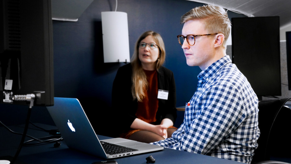 Bilden visar en manlig student sittandes framför en dator. I bakgrunden syns en kvinnlig lärare.