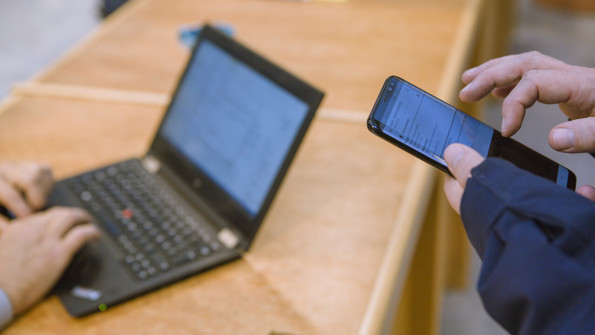 Närbild på person som håller i smartphone och en annan person som knappar på en bärbar dator.