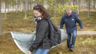 Två personer som bär en kanot genom skogen mot vattnet.