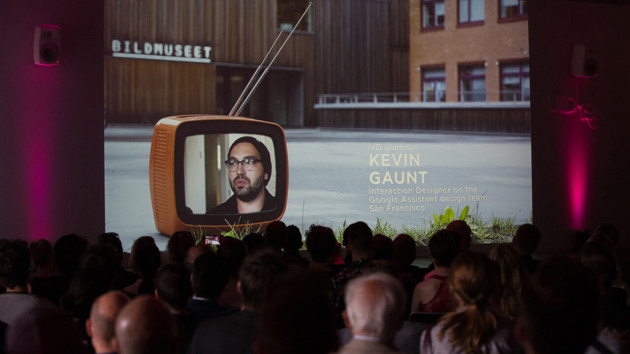 många personer sitter samlade i ett auditorium med ryggarna mot betraktaren. De ser på en stor projicerad bild som föreställer en Tv ståendes på en asfaltsplan. På tv-skärmen syns en ung man med skägg.