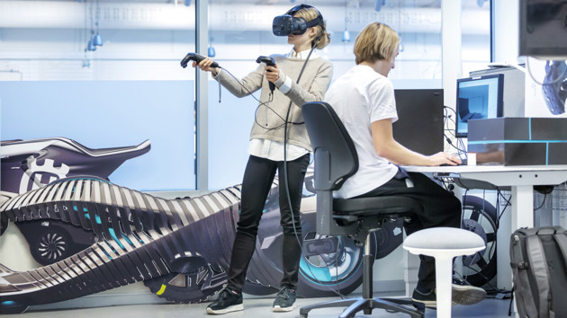 Två studenter visualiserar idéer med hjälp av 3D-moddelering och VR-glasögon. En av dem, en tjej, har VR-glasögon på sig och navigerar med två handkontroller. Den andre, en kille, sitter framför en datorskärm med en 3D-modell på.